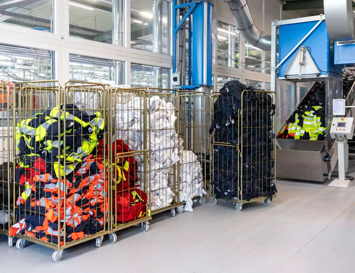 In einer hellen Industriewäscherei mit großen Maschinen und Metallrohren reihen sich Wagen mit gut sichtbarer Arbeitskleidung und weißer Wäsche aneinander.