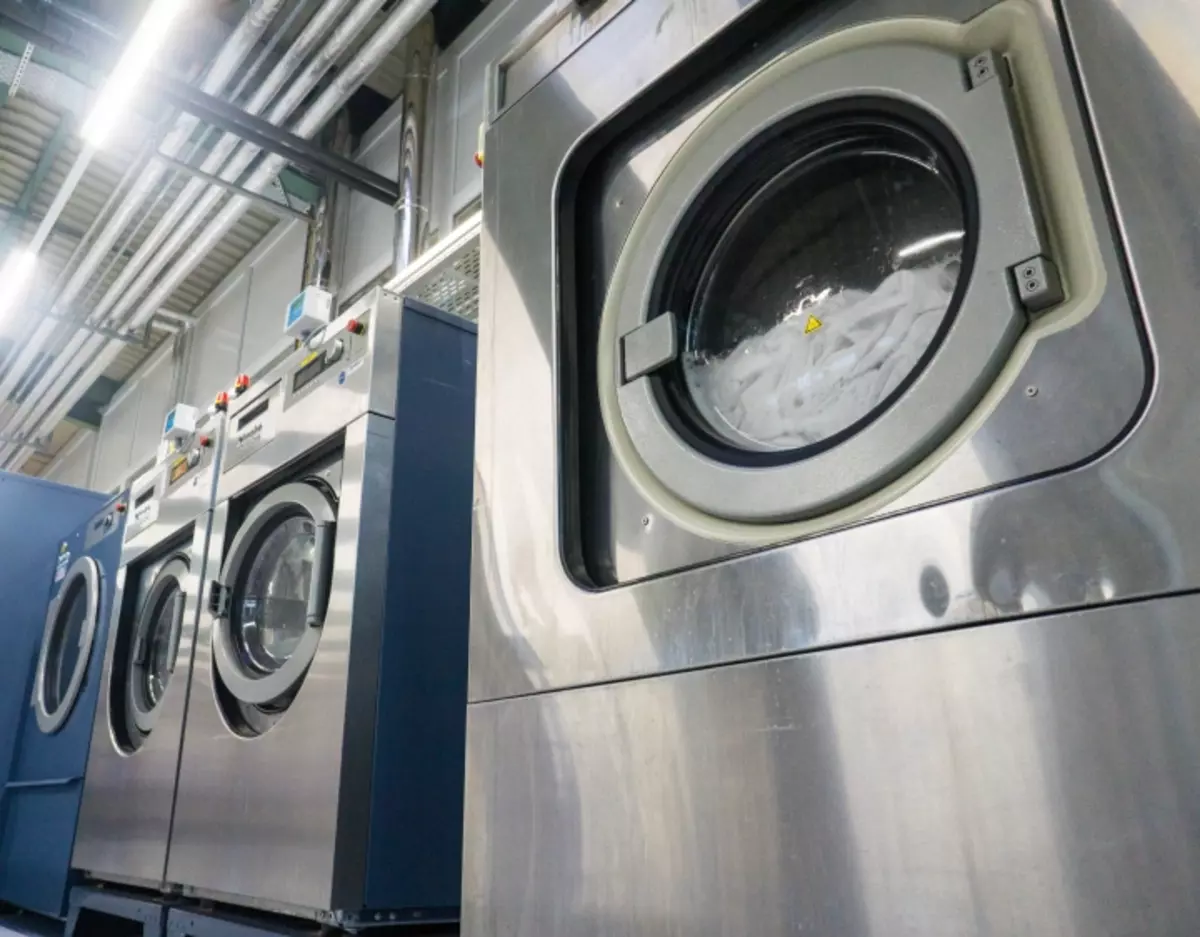 Reihe großer industrieller Waschmaschinen, in denen Arbeitskleidung im Einzelhandel gewaschen wird, mit Edelstahlfronten in einer gewerblichen Wäscherei, einige Maschinen in Betrieb mit sichtbarer Wäsche darin, umgeben von freiliegenden Rohren und einer hohen Decke.