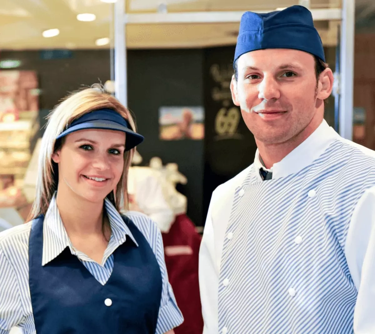 Arbeitskleidung im Einzelhandel: Zwei Bäckereimitarbeiter in blauen Uniformen und Hüten stehen nebeneinander und lächeln in die Kamera in einer hell erleuchteten Bäckerei. Im Hintergrund sind Regale mit Backwaren zu sehen.