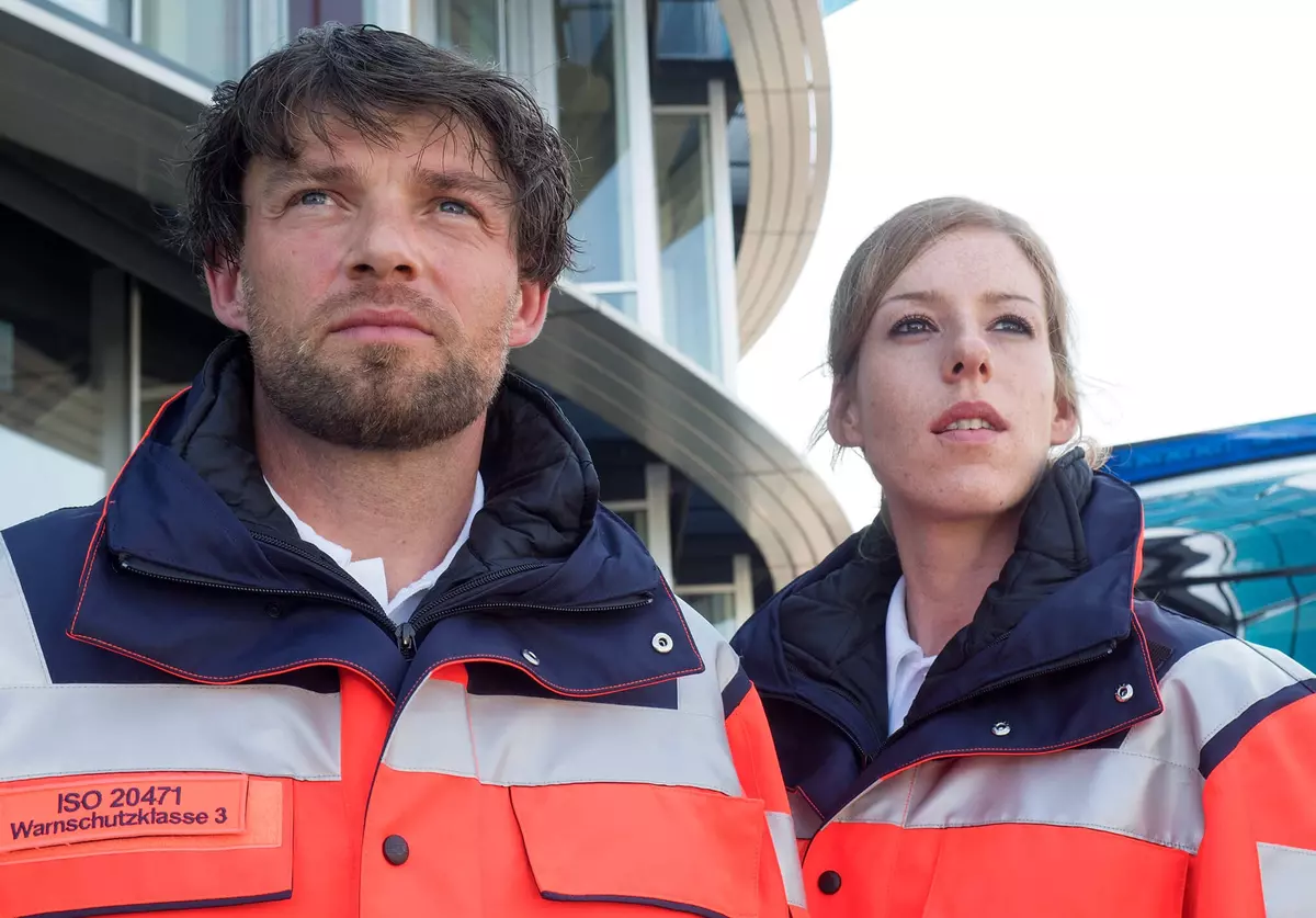 Ein Mann und eine Frau in Warnschutzkleidung beide schauen ernst in die Ferne. Die Kleidung ist blau, weiß und orange. Im Hintergrund ist ein Gebäude zu sehen.