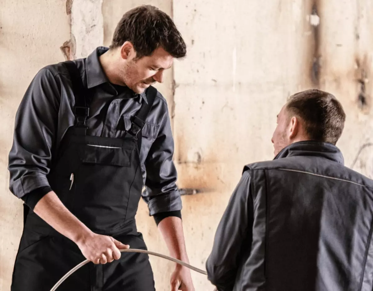 Ein Mann mittleren Alters zieht an einem Kabel. Er ist in Arbeitskleidung bekleidet. Vor ihm steht ein Mann, der zuschaut und ebenfalls Berufsbekleidung trägt.