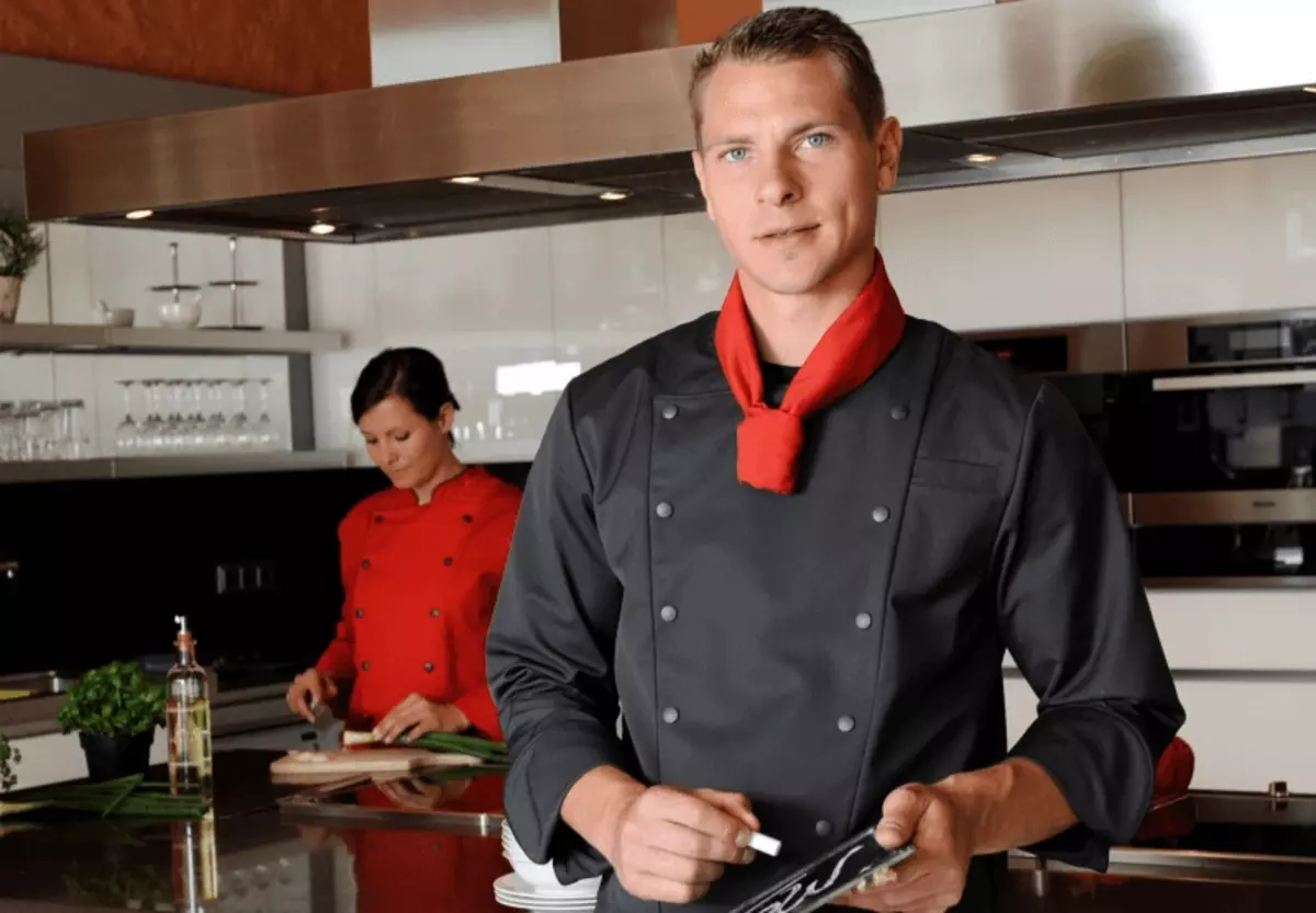Ein blonder Mann mit afel und Kreide in der Hand. Er trägt die Kleidung eines Kochs. Auf der Tafel steht Menü. Im Hintergrund ist eine Küche und eine braunhaarige junge Dame in roter Koch-Kleidung zu sehen. Beide tragen Arbeitskleidung Köln.