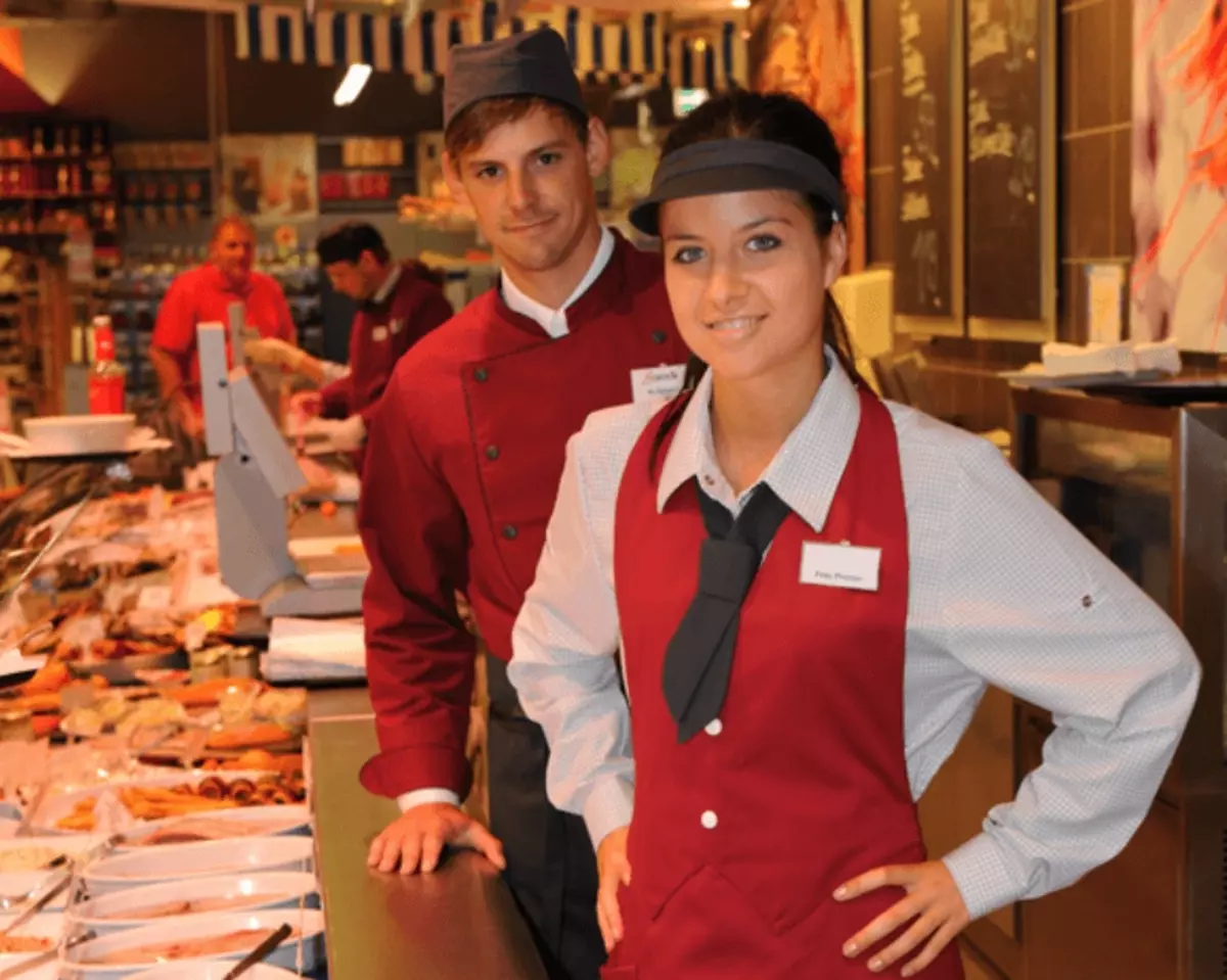 Ein junger Mann mit braunen kurzen Haaren und roter Arbeitskleidung steht lächelnd hinter einer Frau. Auch die blonde Frau mit Arbeitsmütze und Berufsbekleidung in rot-weiß lächelt.