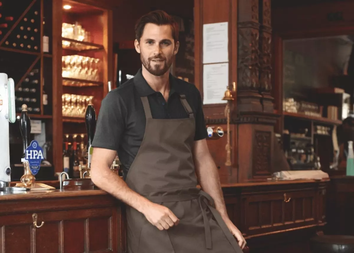 Ein junger Mann mit braunen Haaren steht in Kochkleidung vor einem Thresen. Hinter dem Thresen stehen diverse Flaschen auf einem Regal.